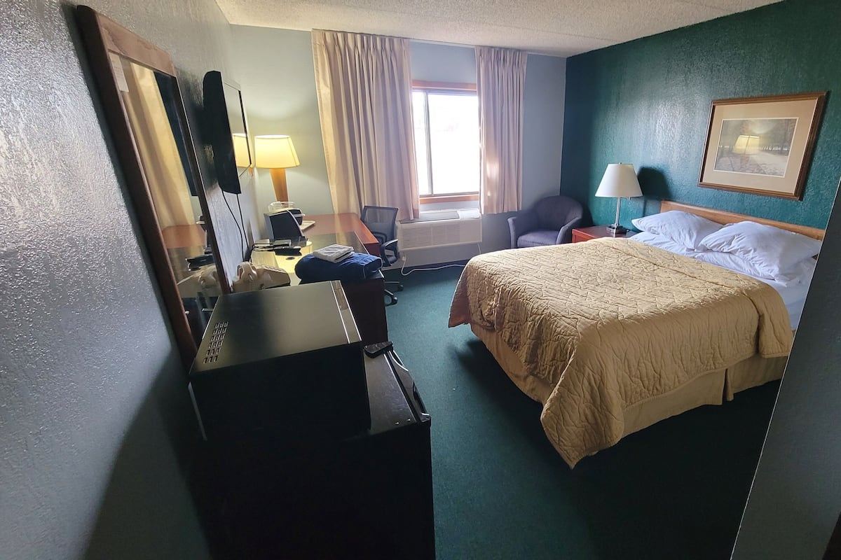 A hotel room with a bed, a desk with a lamp, a chair, a window with curtains, and a framed picture on the wall.