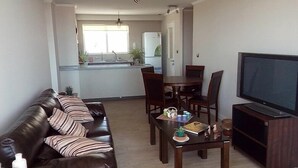 A living room with a dark brown leather sofa, a wooden coffee table with books and cups, a flat-screen TV, and a dining area with a wooden table and chairs.