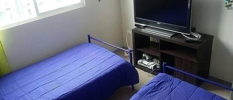 Habitación con dos camas individuales, un televisor sobre un soporte y una ventana con vistas a edificios.