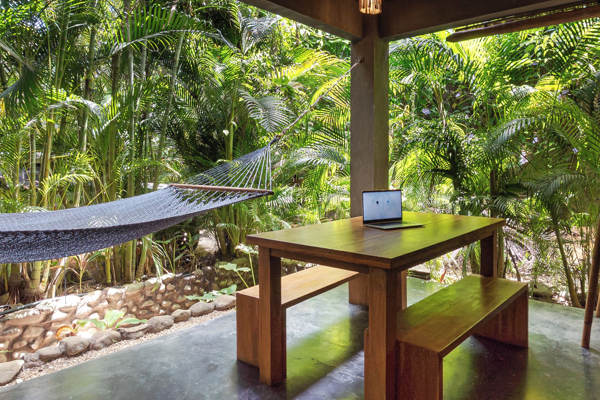 A hammock, a wooden table with a laptop, and lush greenery in a serene outdoor setting.