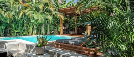 A pool area with a wooden deck, stone steps, and lush tropical plants.