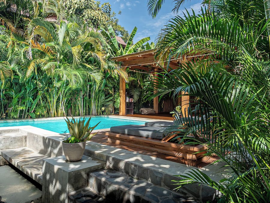 A pool area with a wooden deck, stone steps, and lush tropical plants.