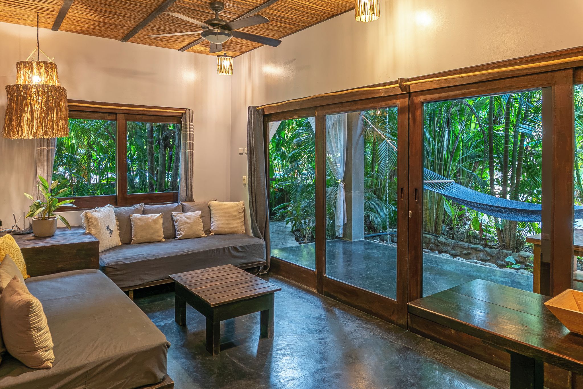 A spacious room with a large sliding glass door leading to a tropical garden, a comfortable seating area with a sofa and a coffee table, and a ceiling fan.