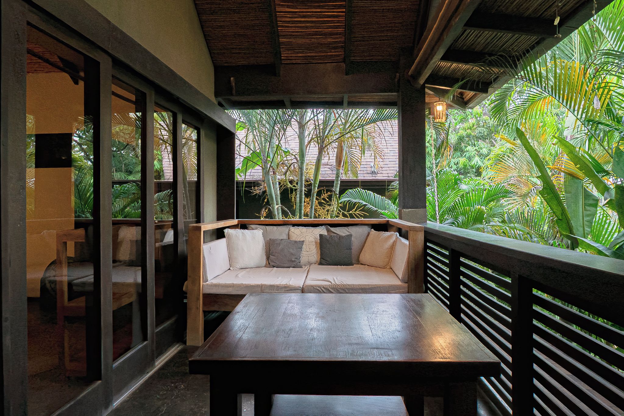 An outdoor seating area with a wooden table and sofa, surrounded by glass walls and a view of tropical vegetation.
