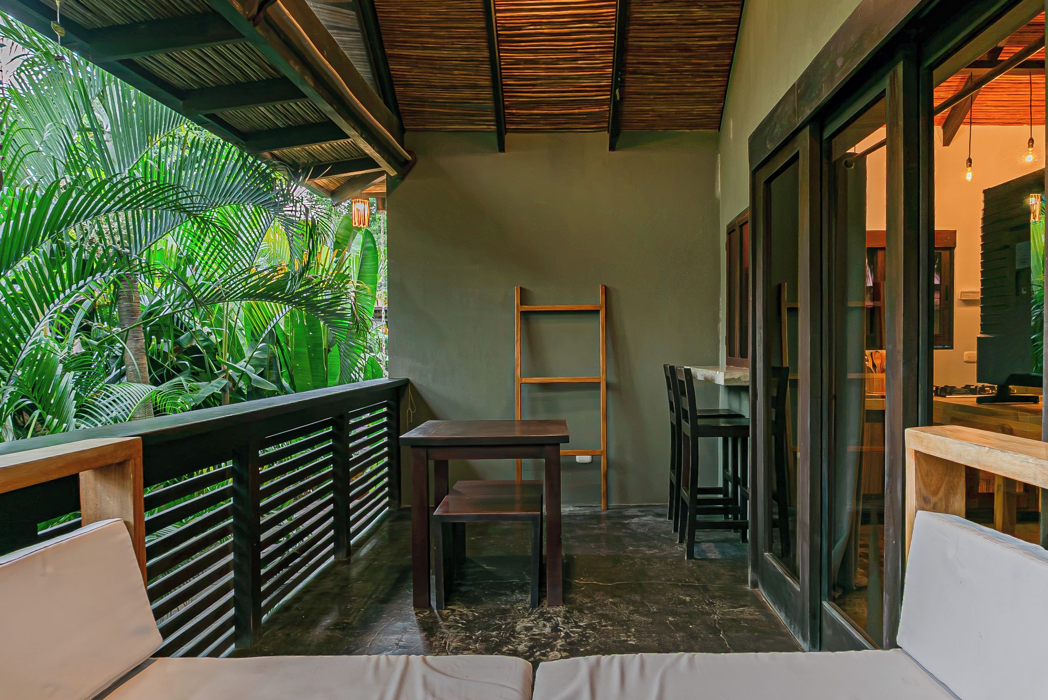 A balcony with a wooden table and chairs, a small wooden ladder, and a view of lush green foliage.