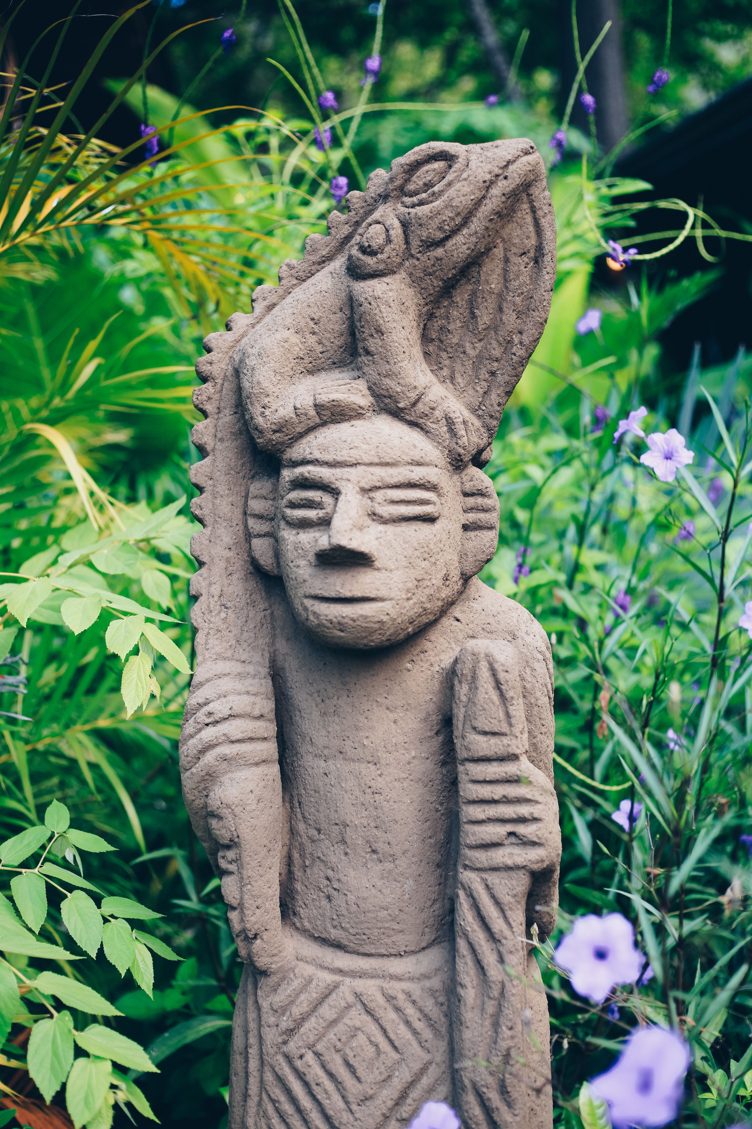 A stone statue in a garden with green foliage and purple flowers.