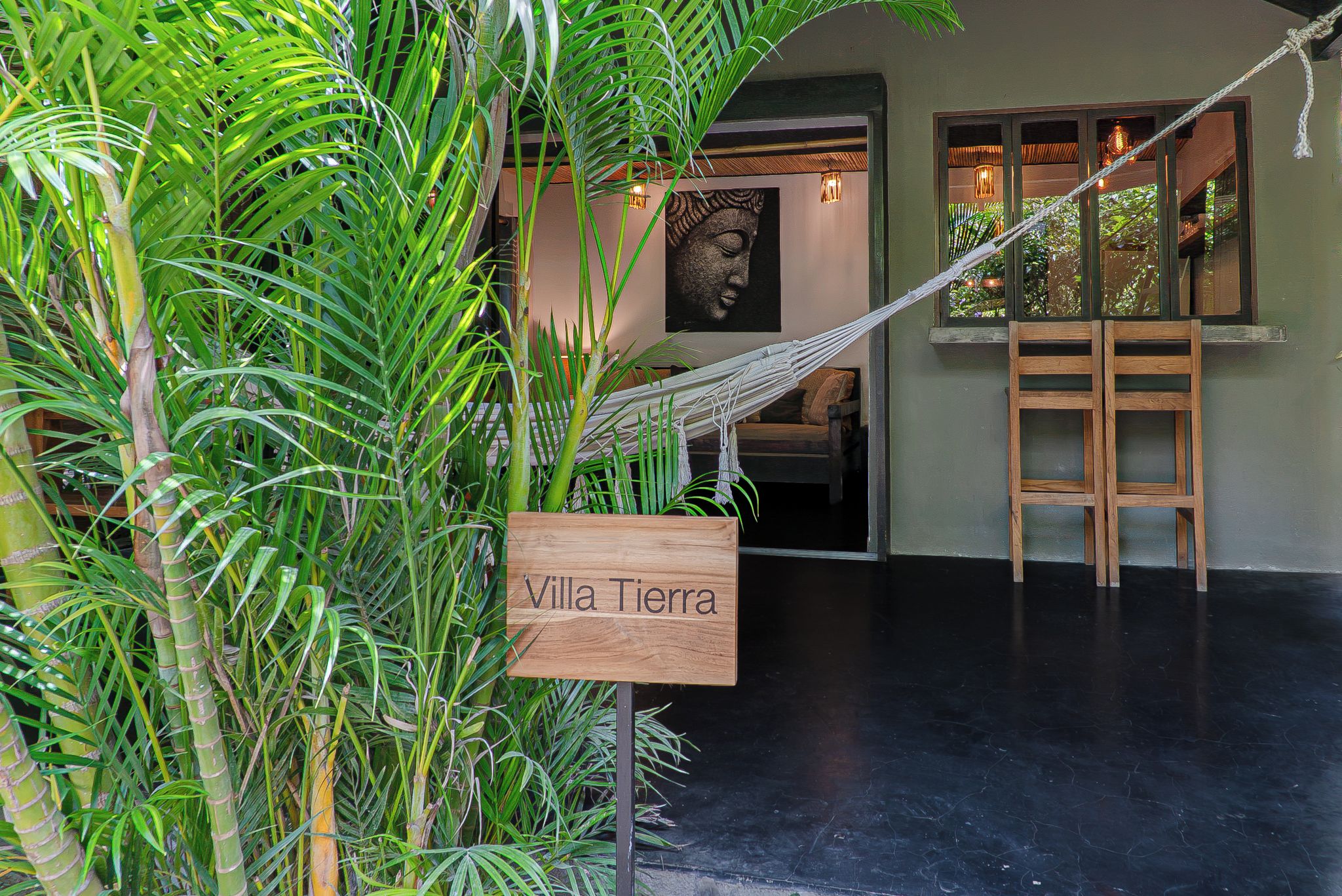 A room with a hammock, a wooden sign reading 'Villa Tierra', and a large window with a view of greenery.