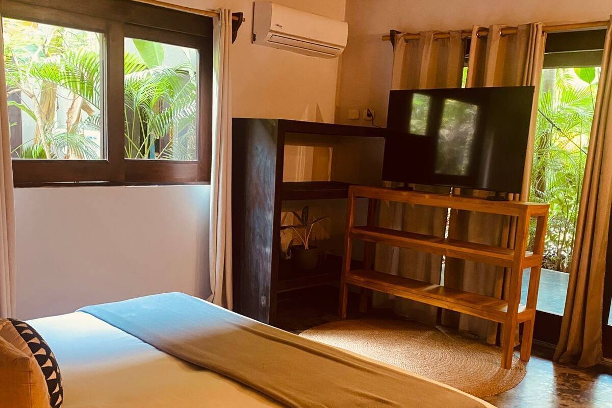 A bedroom with a bed, a TV on a wooden stand, and a window with a view of greenery.