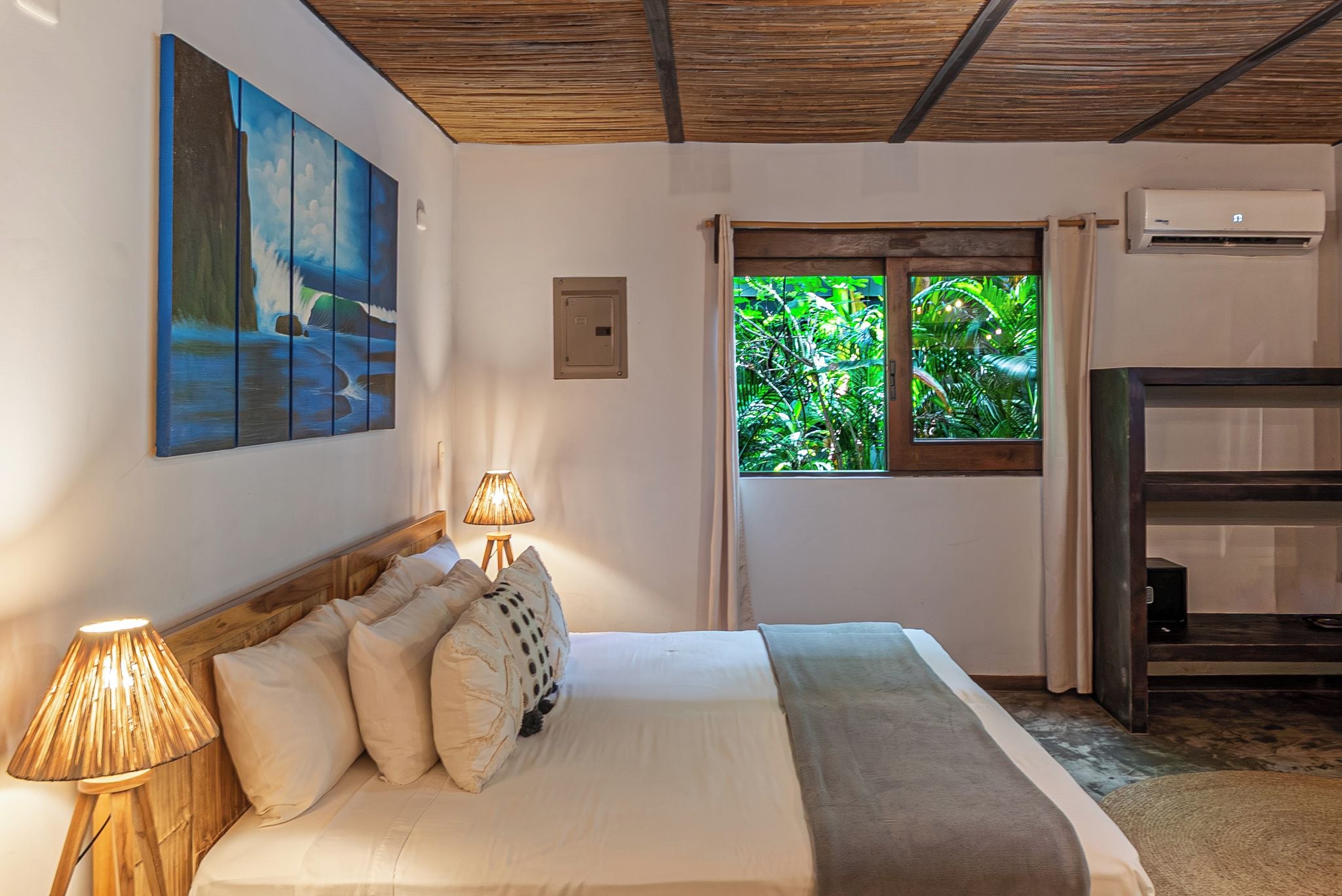 A bedroom with a bed, pillows, a lamp, a painting, a window with a view of greenery, and an air conditioning unit.