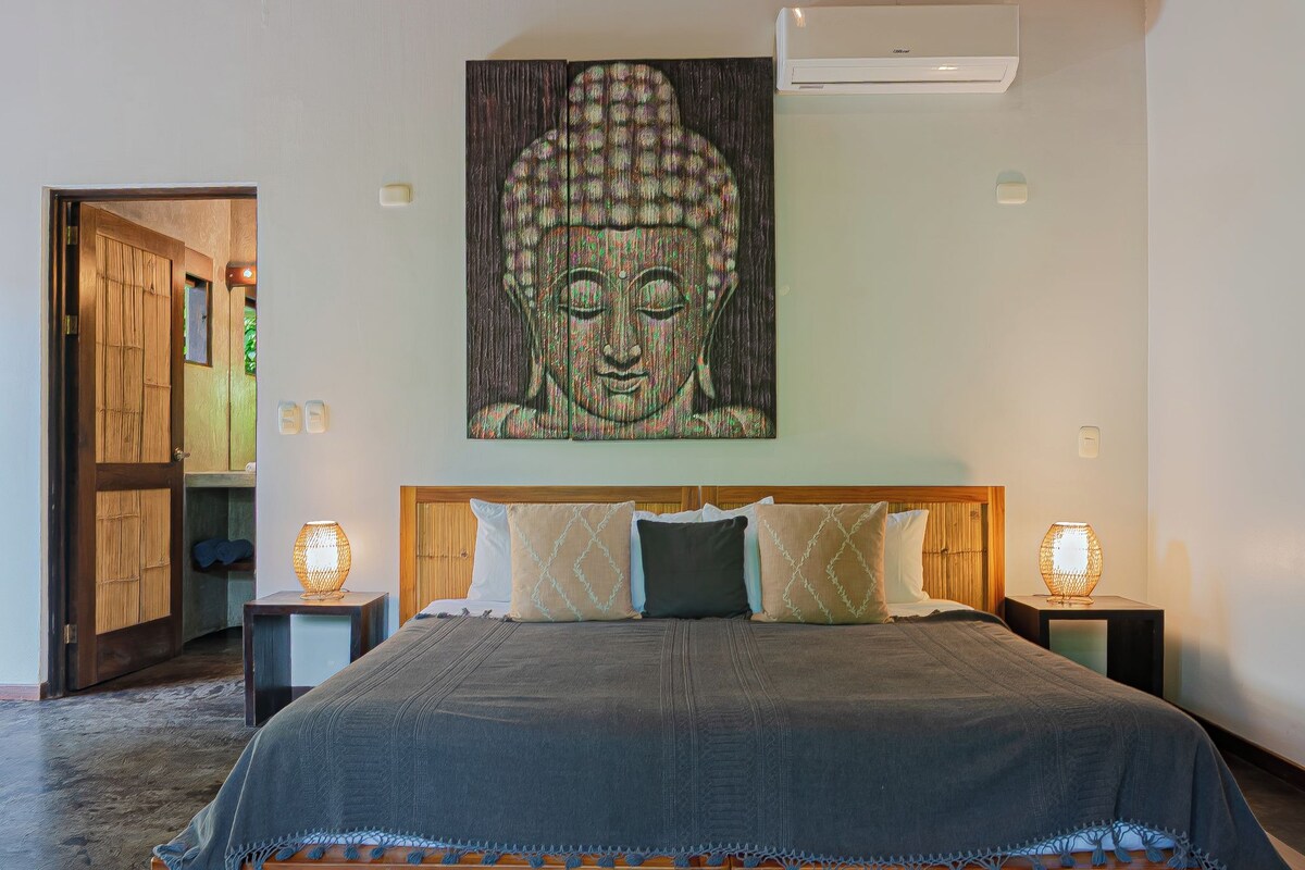 A bedroom with a large bed, two bedside tables with lamps, a wall-mounted air conditioner, and a wooden door.