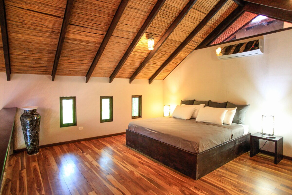 A bedroom with a wooden ceiling, a large bed, and a vase on the floor.
