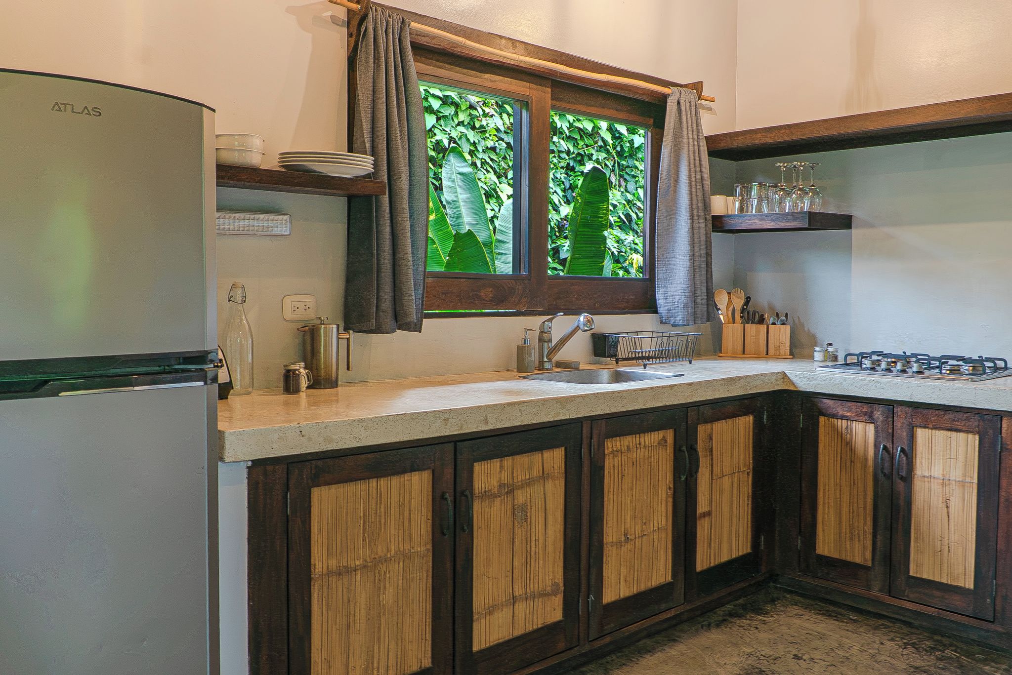 A kitchen with a refrigerator, sink, wooden cabinets, and a window with curtains.
