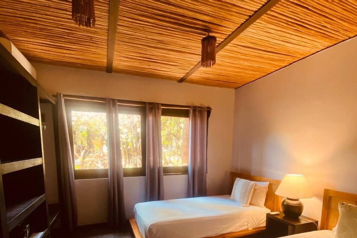 A room with two beds, a wooden ceiling, a window with curtains, and a nightstand with a lamp.