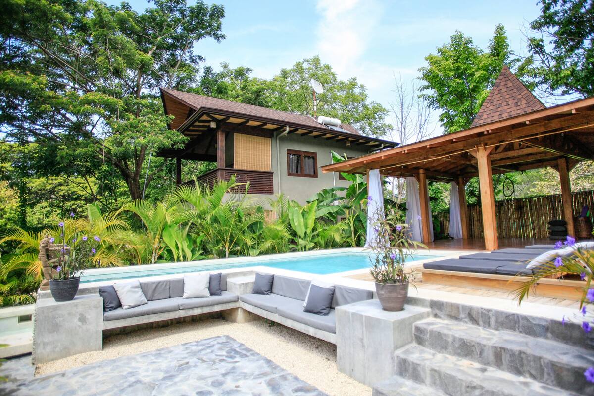 A poolside area with a built-in seating area, a covered patio, and lush greenery.