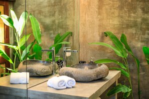 A bathroom with a stone sink, two faucets, and a mirror. There are towels on the counter and green plants around.