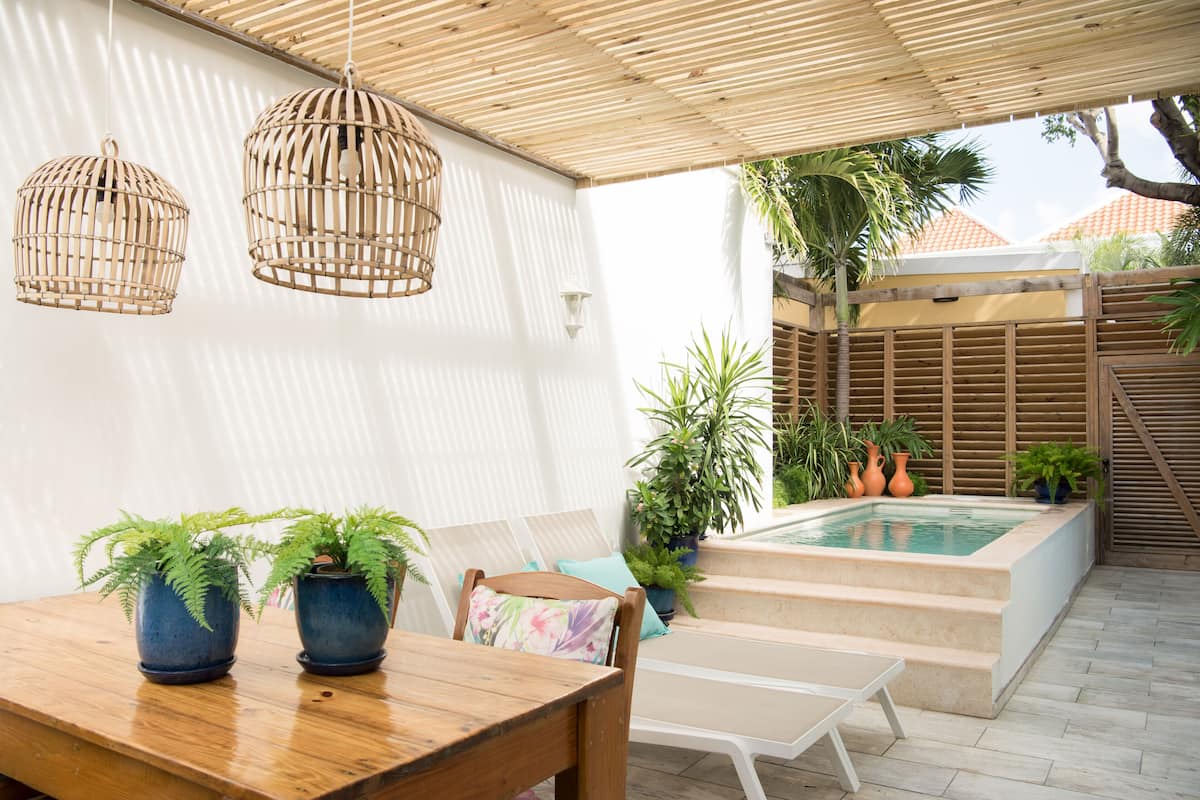 Un patio con una mesa de madera, dos lámparas colgantes de mimbre, una piscina y plantas en macetas.