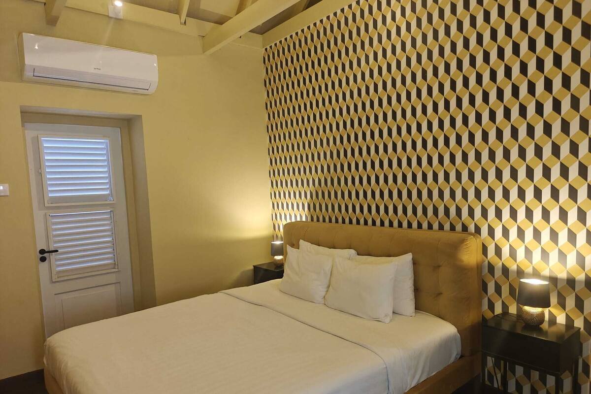 Una habitación de hotel con cama, una puerta con persianas, un papel tapiz estampado y una mesita de noche con lámpara.