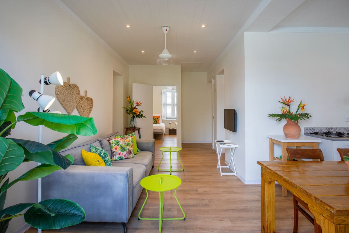 Una sala moderna con un sofá gris, cojines verdes y amarillos, una pequeña mesa verde y una mesa de comedor de madera.