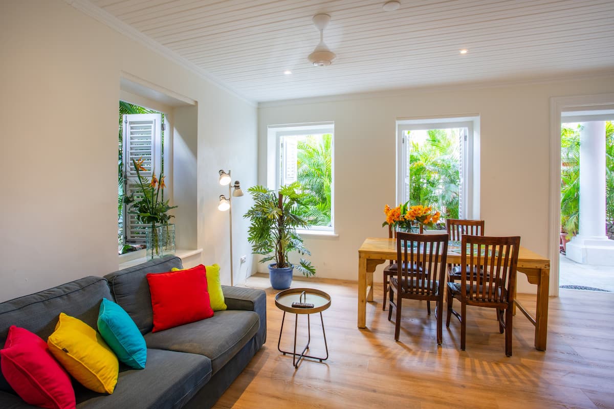 Una sala de estar con un sofá gris, cojines coloridos, una mesa de comedor de madera con sillas, una planta en maceta y ventanas amplias con vistas a zonas verdes.