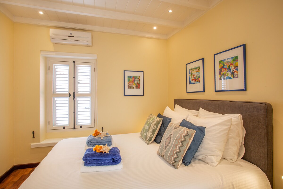 Une chambre avec un grand lit, une fenêtre avec des volets et deux tableaux encadrés accrochés au mur.