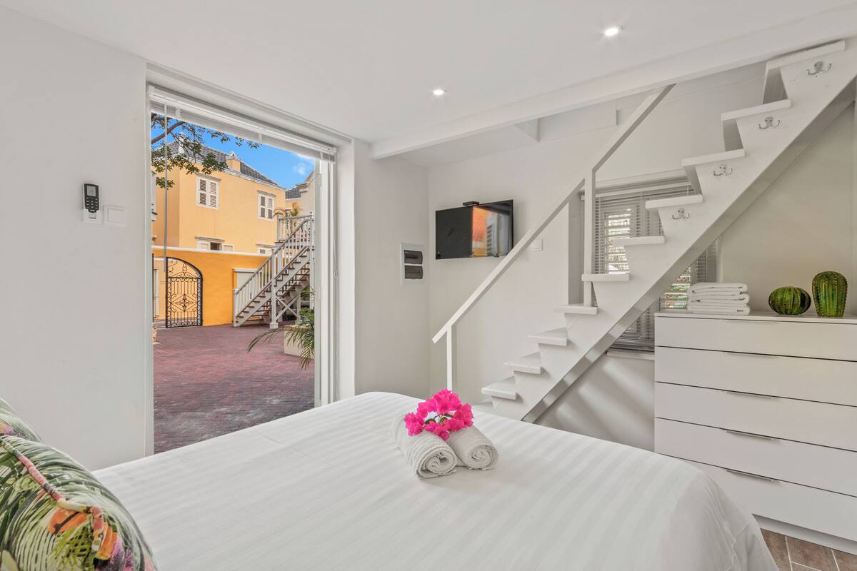 Habitación de hotel con cama, una escalera blanca, televisión y vista a un edificio amarillo exterior.