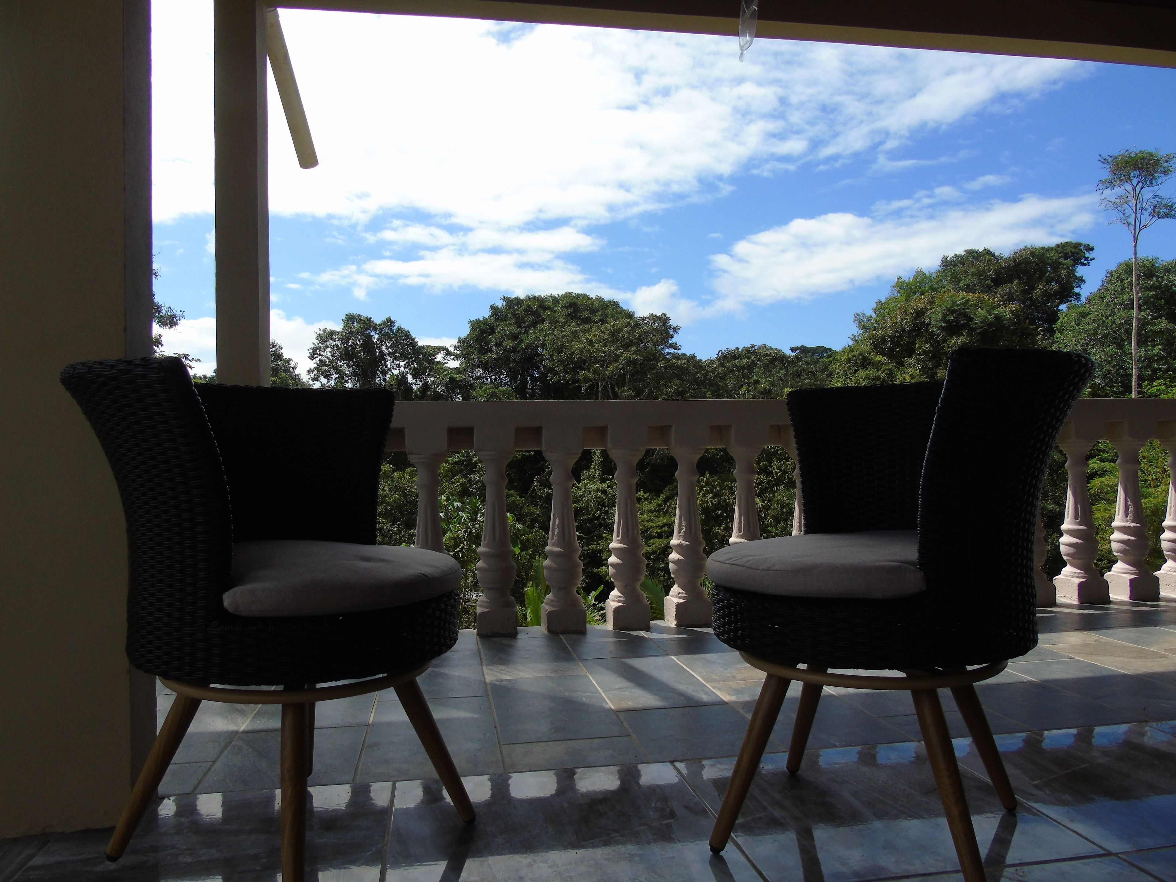 Two wicker chairs with wooden legs on a tiled balcony with a balustrade, overlooking a lush green landscape and a partly cloudy sky.