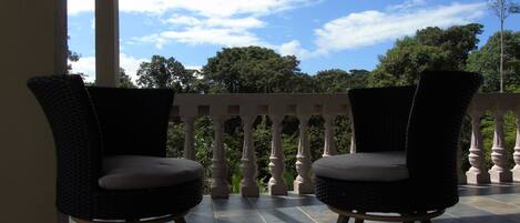 Two wicker chairs with wooden legs on a tiled balcony with a balustrade, overlooking a lush green landscape and a partly cloudy sky.