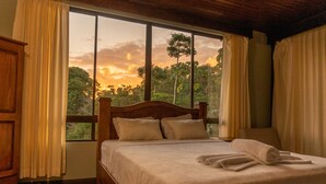 A bedroom with a large window, a wooden bed, and a view of trees and sky. - Hotel Margarita (Drake Bay)