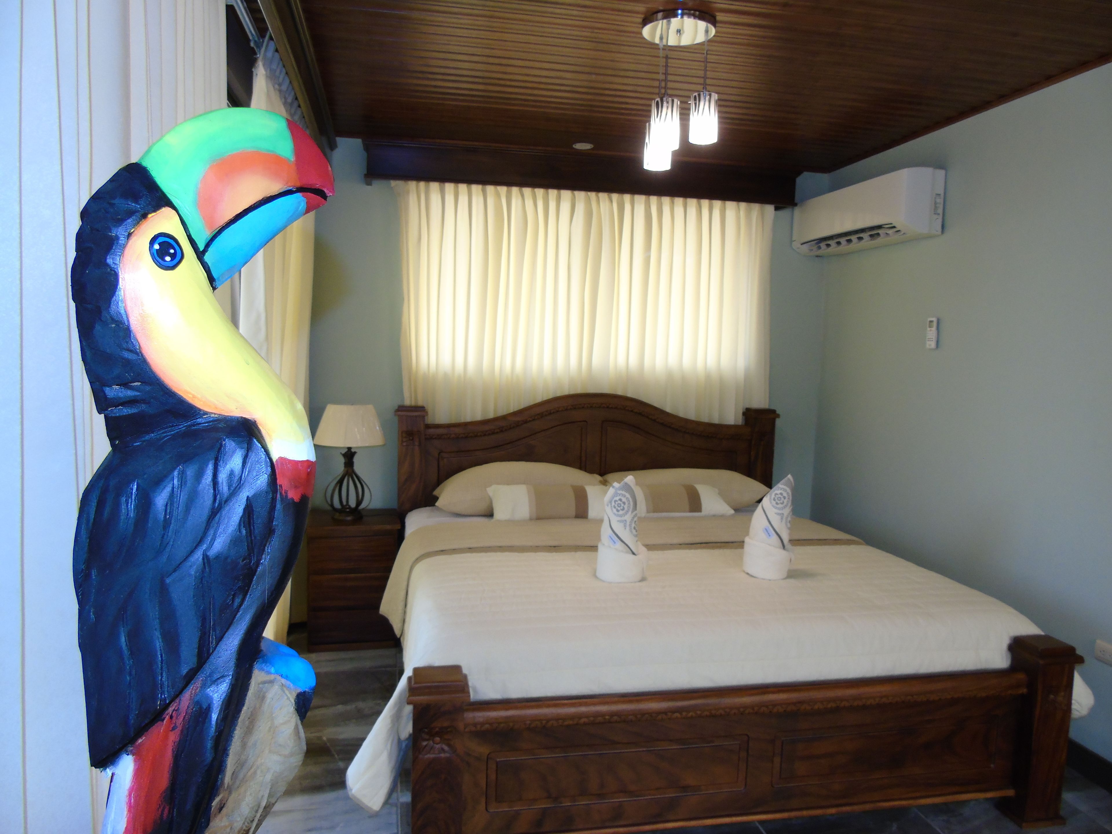 Ein Hotelzimmer mit einem hölzernen Bett, einem Nachttisch mit Lampe, einer großen, farbenfrohen Vogel-Skulptur und einer Deckenleuchte.