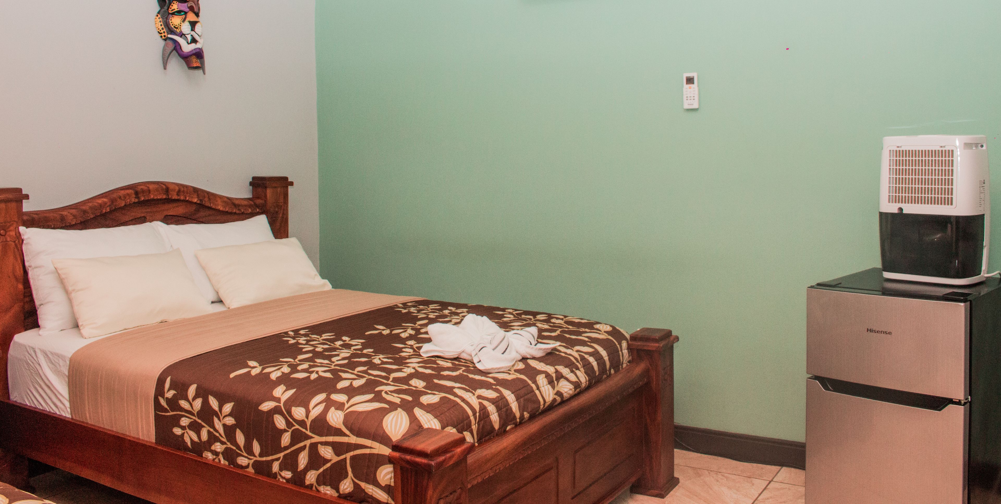 A neatly made bed with a floral bedspread and white pillows, a wooden headboard, a small refrigerator, and a wall-mounted air conditioner.