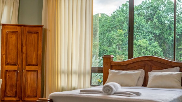 A wooden bed with white linens and pillows, a wooden wardrobe, and a window with a view of dense green foliage. - Hotel Margarita (Drake Bay)
