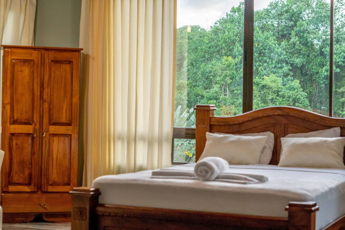 A bedroom with a wooden bed, white bedding, a window with a view of greenery, and a wooden wardrobe.