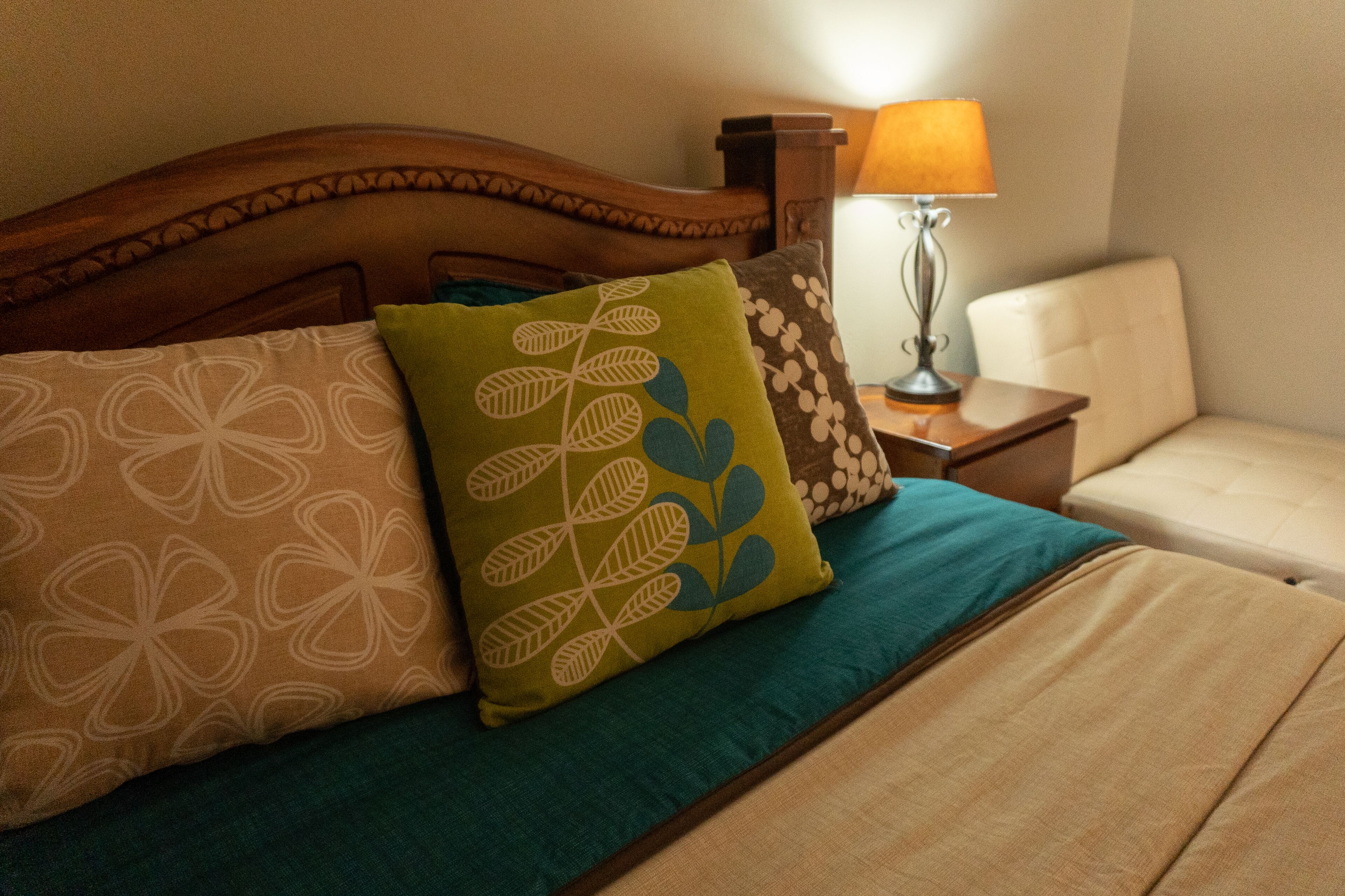 A neatly made bed with a wooden headboard, multiple pillows with floral patterns, a teal bedspread, and a bedside lamp on a nightstand.