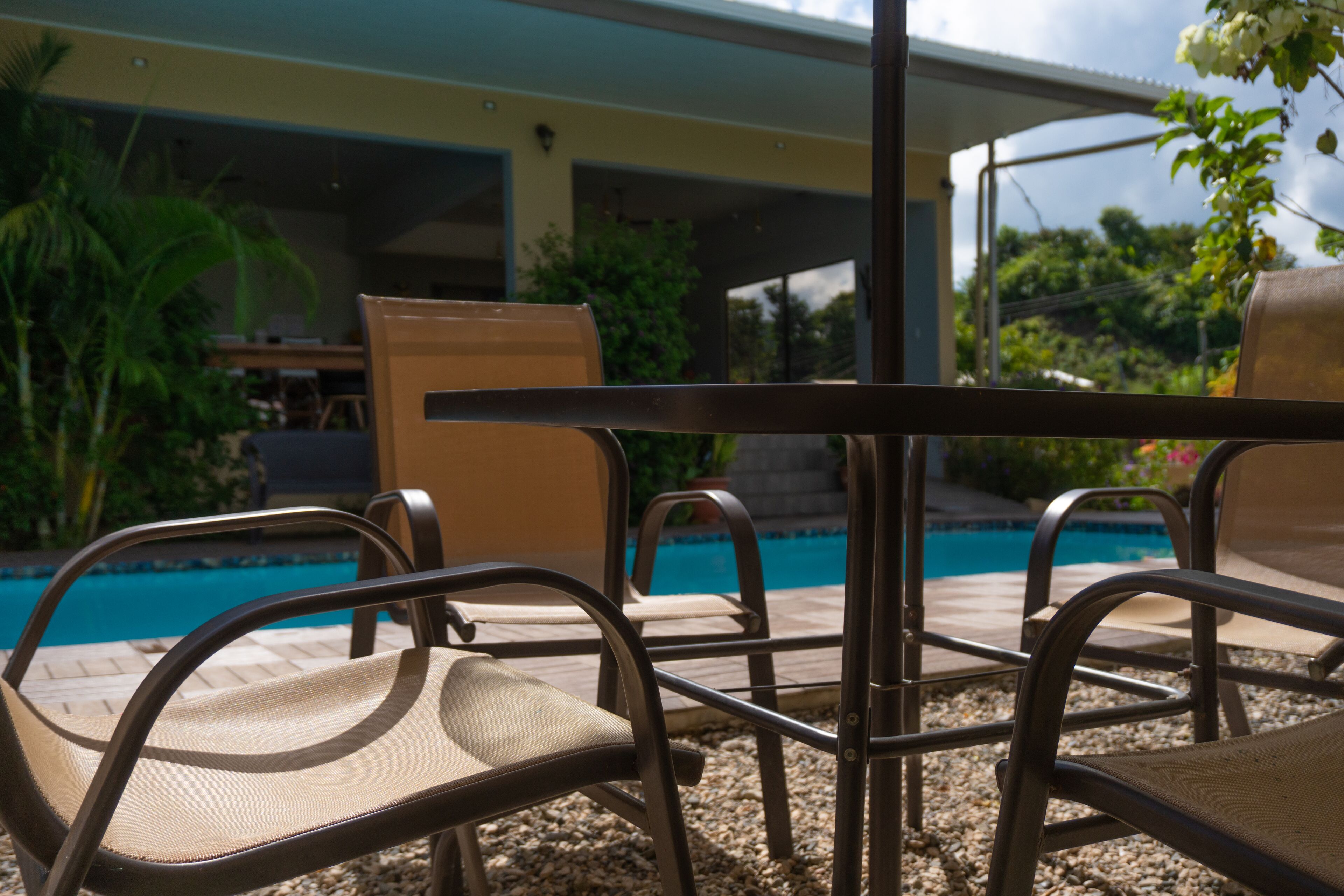 A poolside area with chairs and a table, surrounded by greenery and a building in the background.