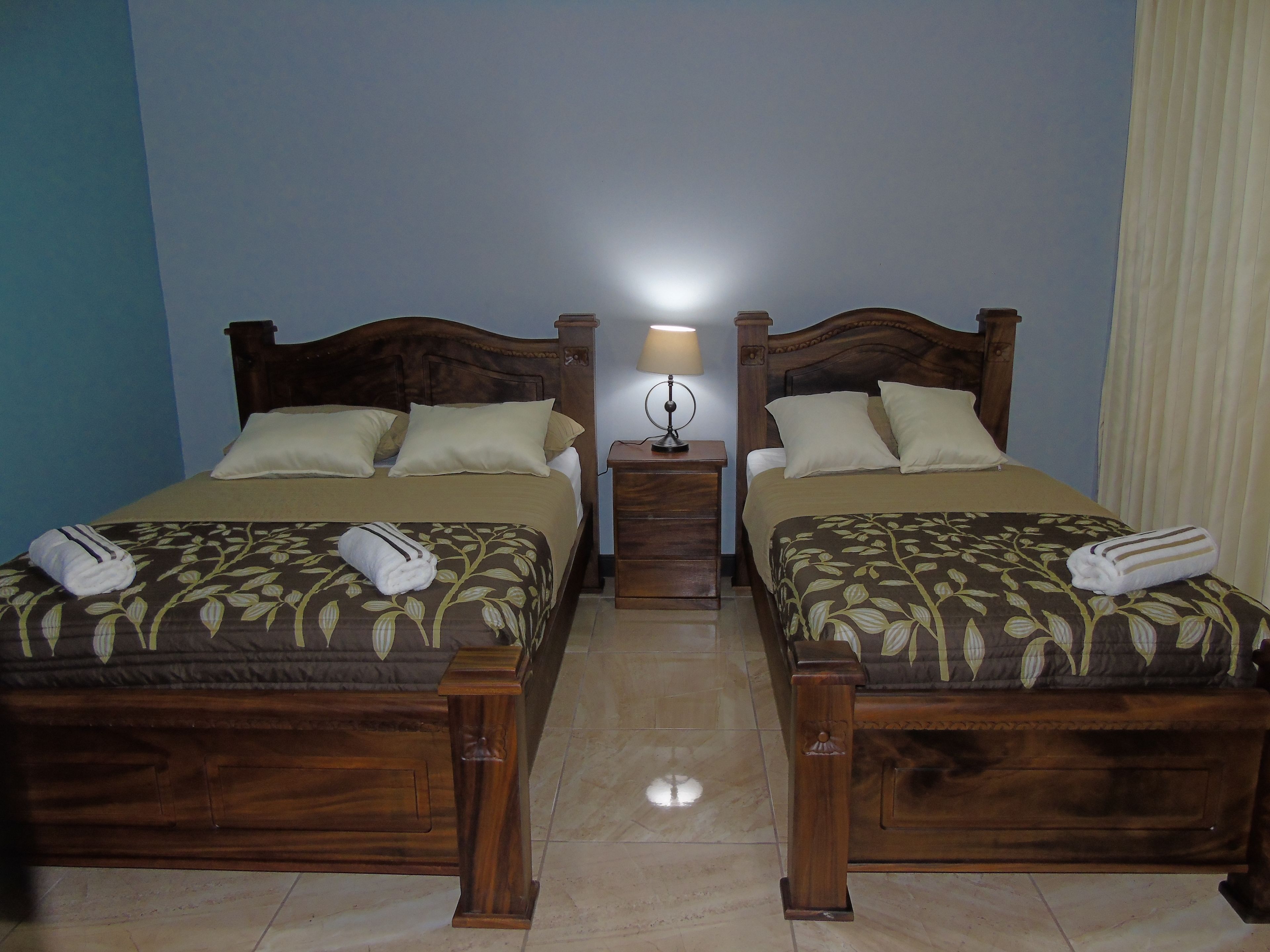 A hotel room with two wooden beds, each with a floral bedspread and pillows, a nightstand with a lamp, and a window with curtains.
