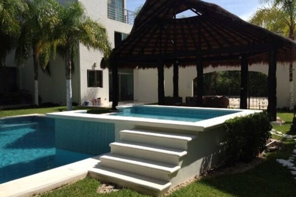 Una piscina con escalones de acceso, rodeada por un edificio blanco y un pabellón con techo de paja.