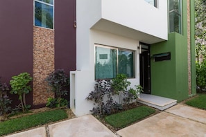 Une maison moderne à deux étages, dotée d’une façade blanche et verte, d’un mur d’accent en briques et d’un petit jardin à l’avant, agrémenté de diverses plantes.