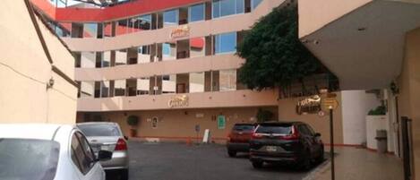 Un hotel de varios pisos con fachada roja y color crema, un estacionamiento con varios automóviles y un letrero que indica el nombre del hotel.