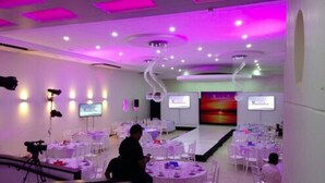 Un salón de conferencias con mesas y sillas redondas, un escenario con una pantalla de presentación e iluminación empotrada en el techo.