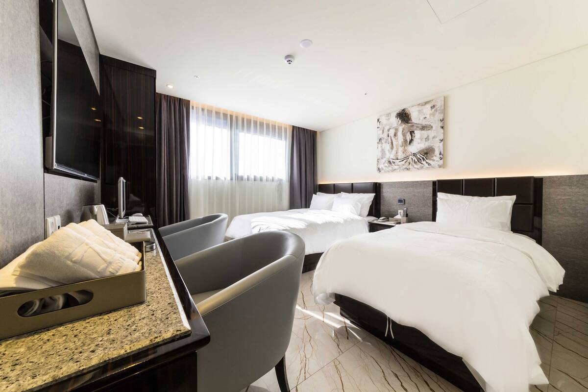 Una habitación de hotel moderna con dos camas, un escritorio y un amplio ventanal con cortinas.