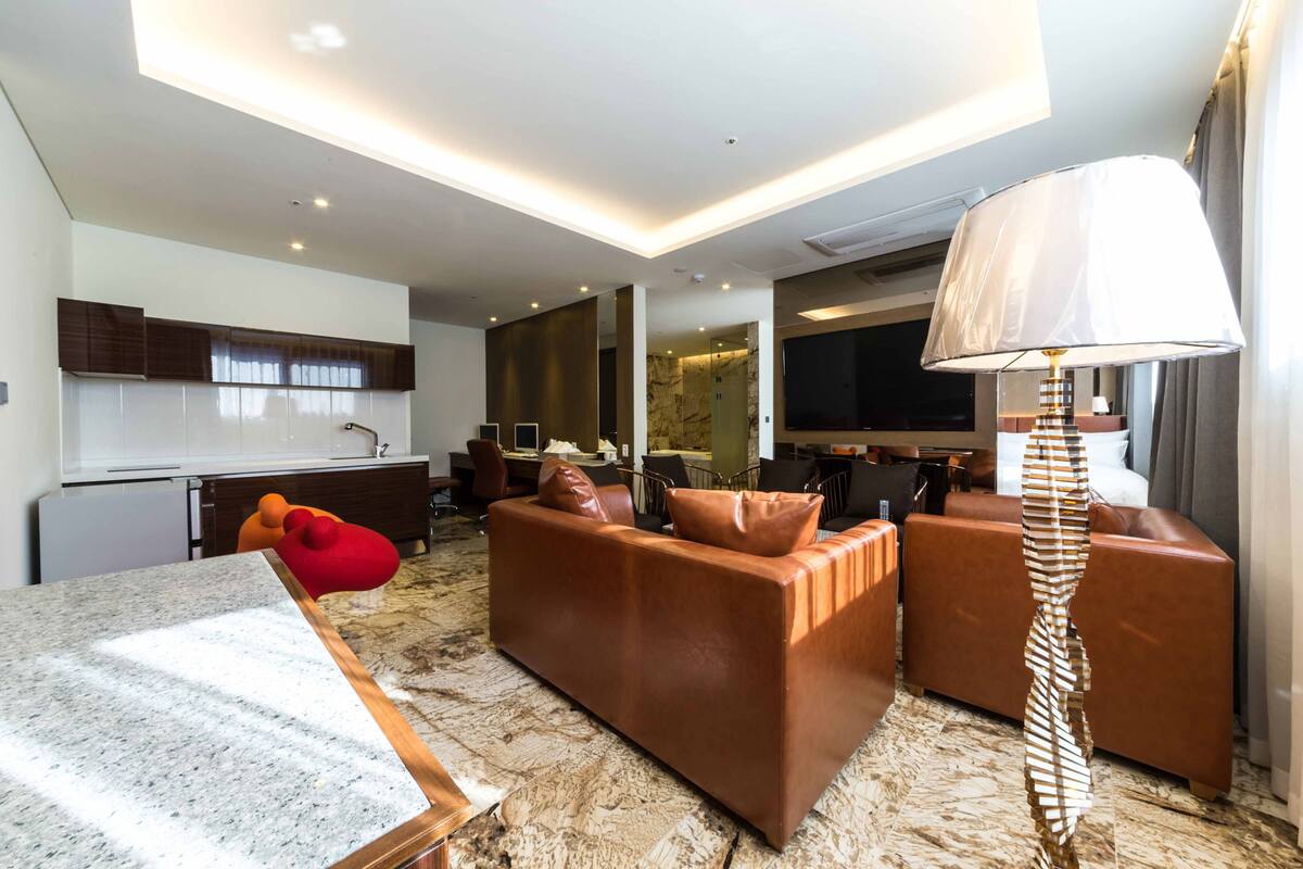 Una habitación de hotel moderna con piso de mármol, sofás de cuero, una zona de comedor y un televisor de pantalla plana grande.