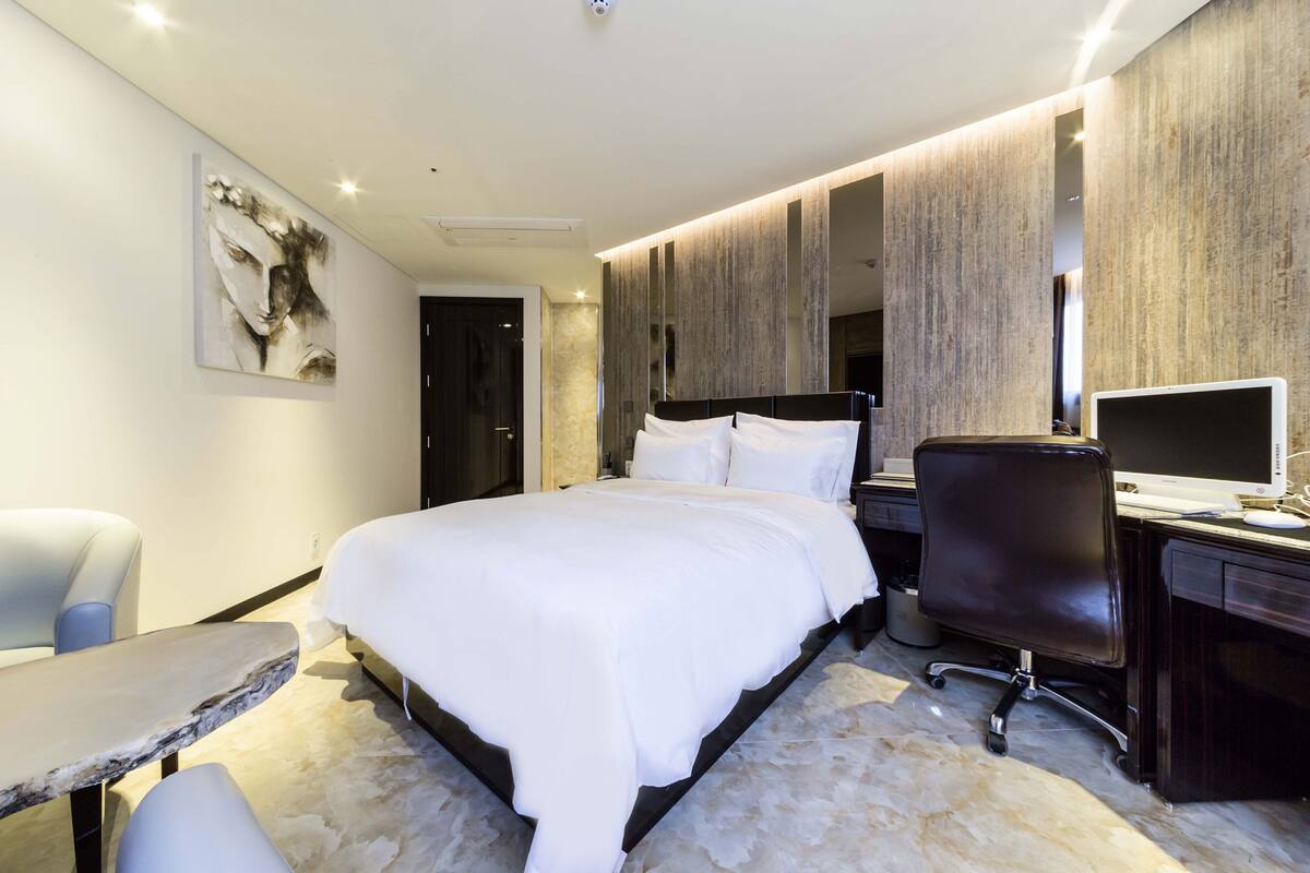 Una habitación de hotel moderna con una cama grande, un escritorio con computadora, una silla y un cuadro en la pared.