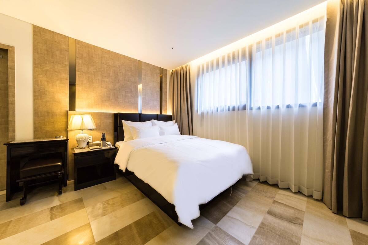 Habitación de hotel con una cama grande, una mesita de noche con lámpara y vistas al exterior a través de cortinas traslúcidas.