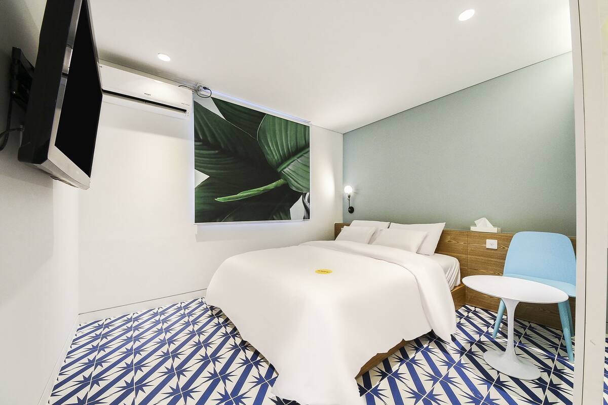 Ein modernes Hotelzimmer mit einem Bett, einem kleinen Tisch, einem Sessel, einem an der Wand montierten Fernseher und einem großen Fenster mit Blick auf Pflanzen.