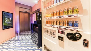 Máquina expendedora con diversas bebidas, una pantalla digital mostrando 'GRAND OPEN' y un piso con diseño.