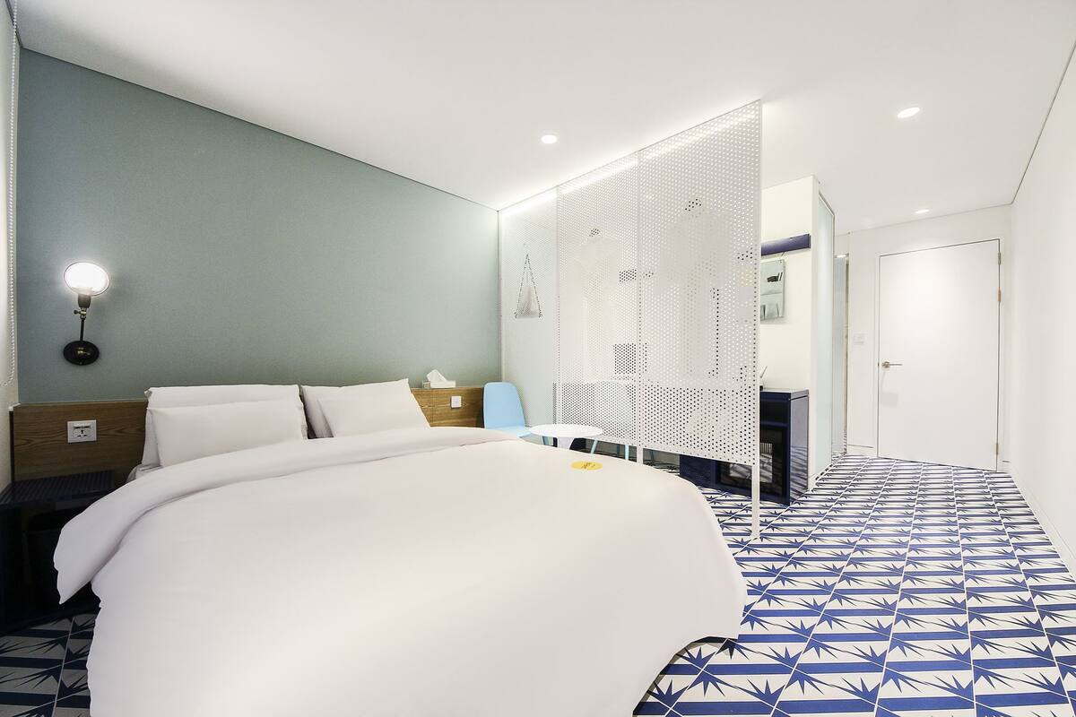 Ein modernes Hotelzimmer mit einem großen Bett, einem Nachttisch, einem Schreibtisch und einem durch eine Trennwand sichtbaren Badezimmer.
