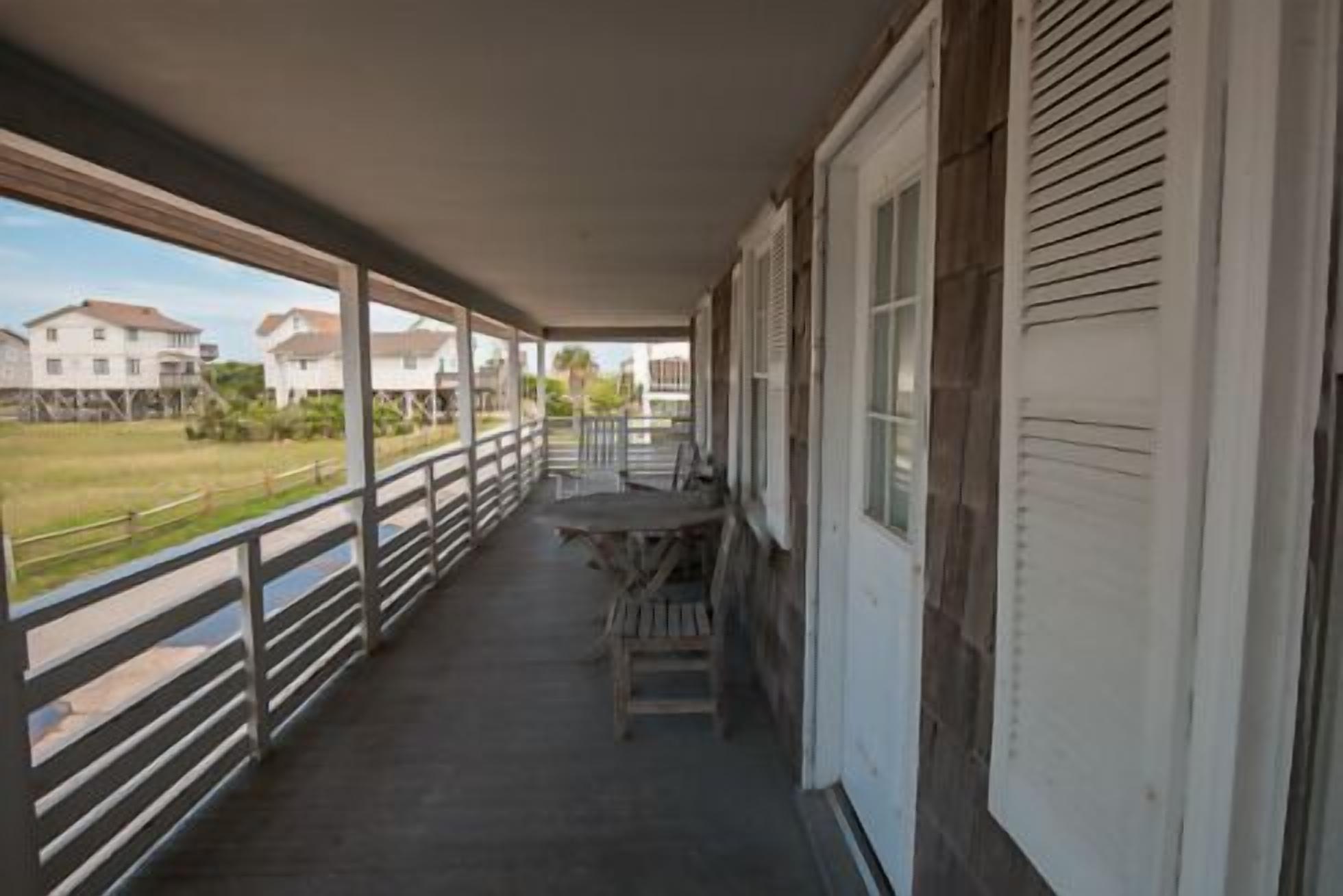 A covered balcony with a table and chairs, overlooking a grassy area and buildings.