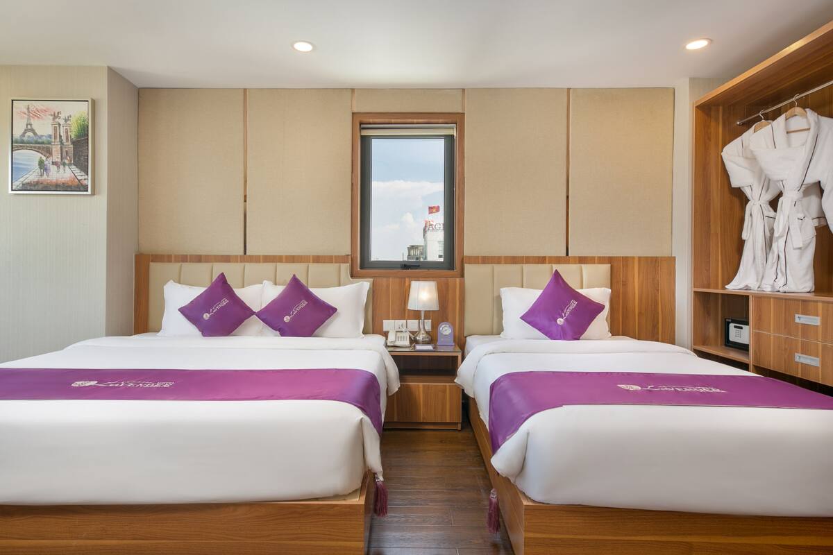 A hotel room with two beds, a wooden wardrobe, a nightstand with a lamp, and a window with a view of a building.