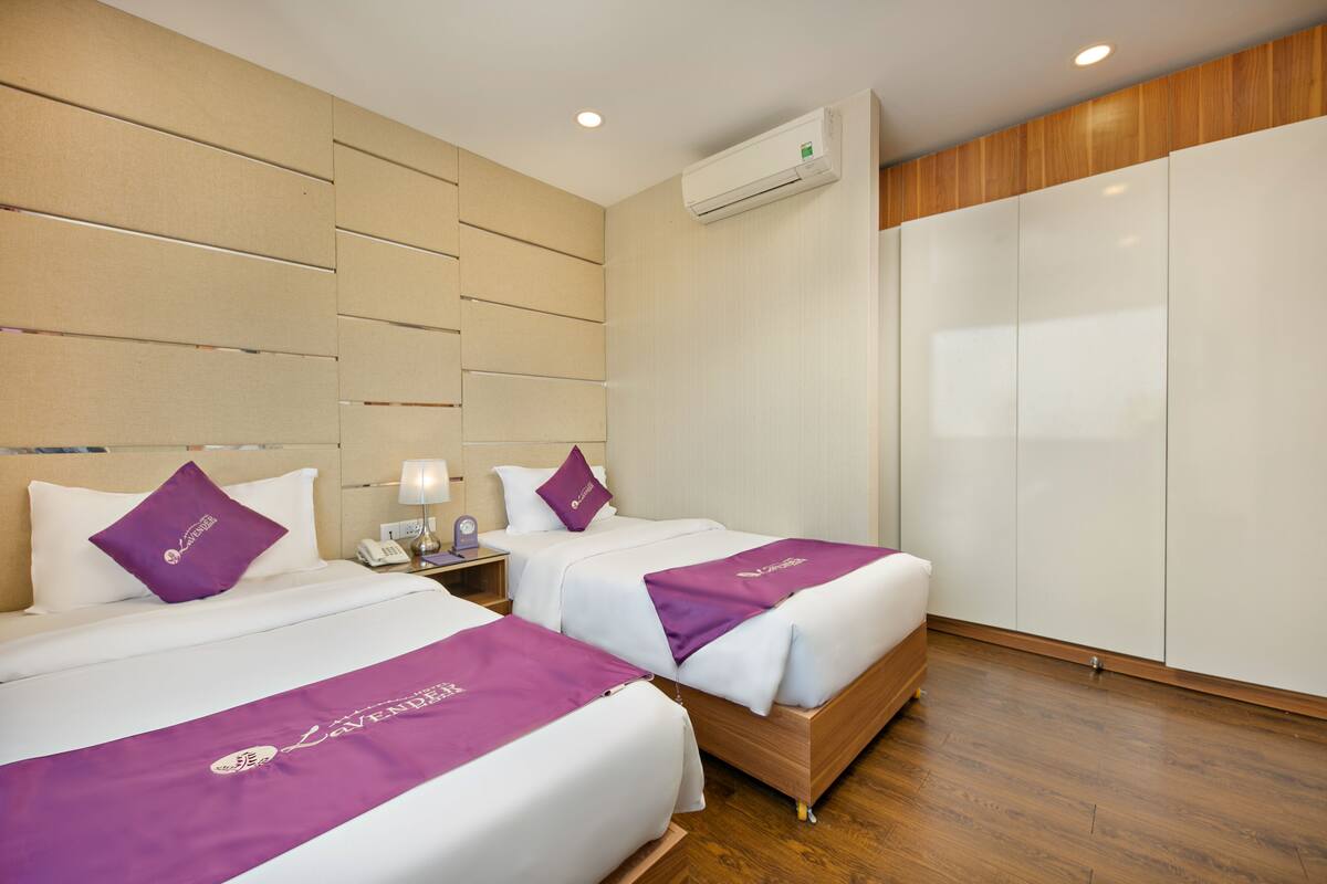 A hotel room with two beds, each with a purple bedspread and matching pillow. There is a wooden floor, a large wardrobe, and an air conditioning unit on the wall.