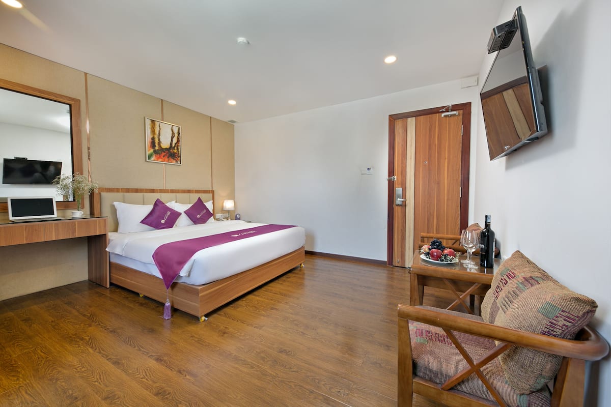 A modern hotel room with a wooden bed, a desk with a laptop, a TV mounted on the wall, and a dining table with a chair.